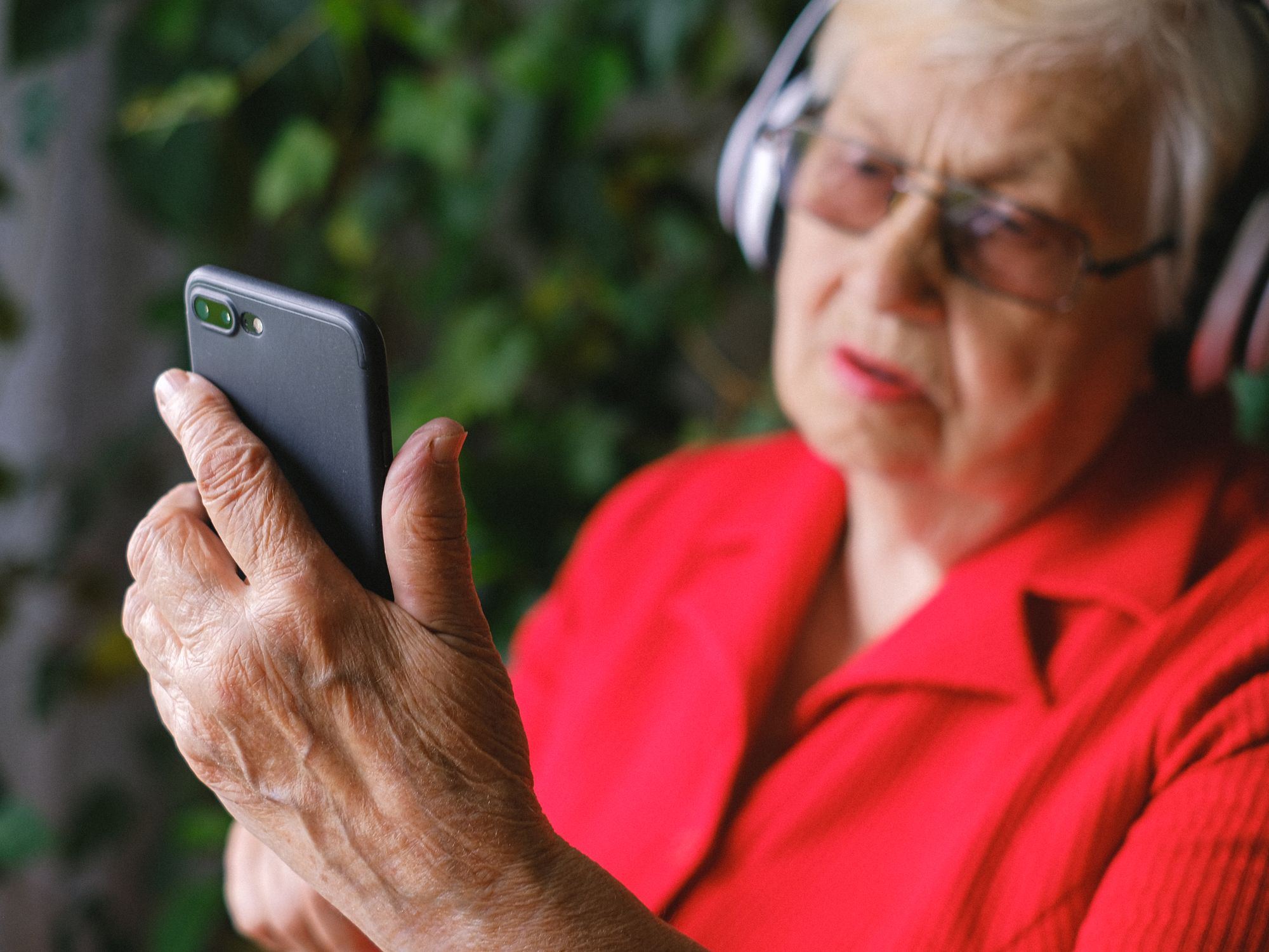 An older woman outdoors looks at her smartphone while wearing headphones.